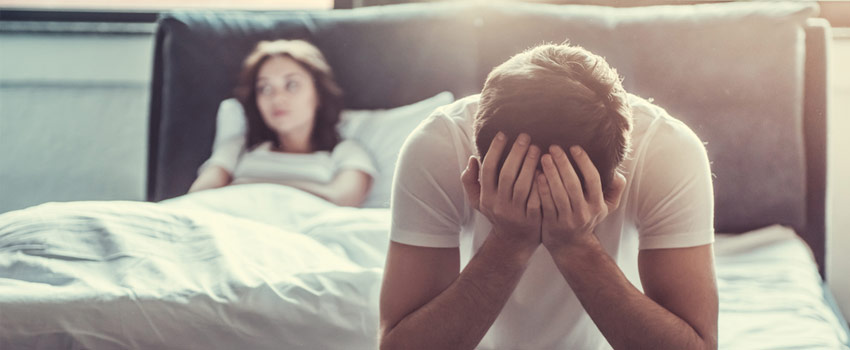 Hombre triste al borde de la cama, con una mujer enfadada a su espalda.